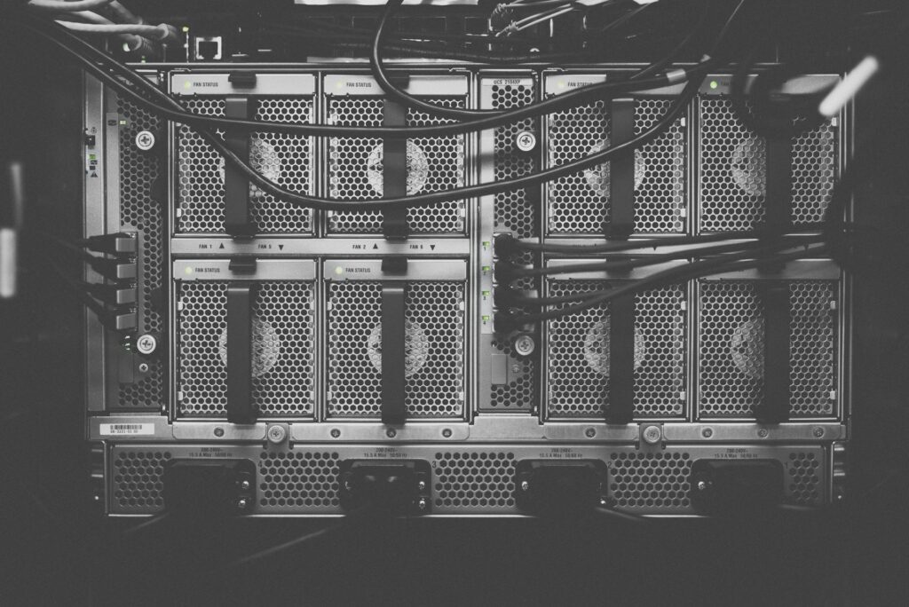 A close-up, top-down view of a server grid with multiple modules, featuring perforated metal panels, ventilation fans, and thick cables, all in a grayscale tone emphasizing the industrial technology.